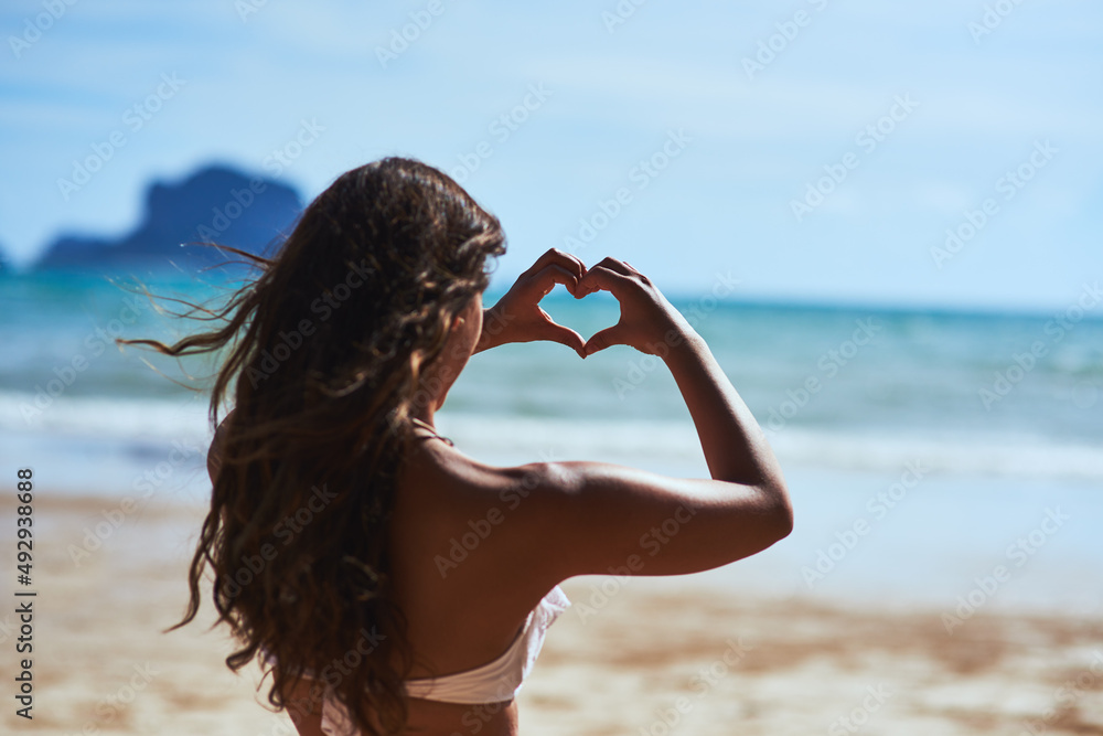 I just love the beach. Shot of a young woman shaping a heart while looking at the scenery at the bea