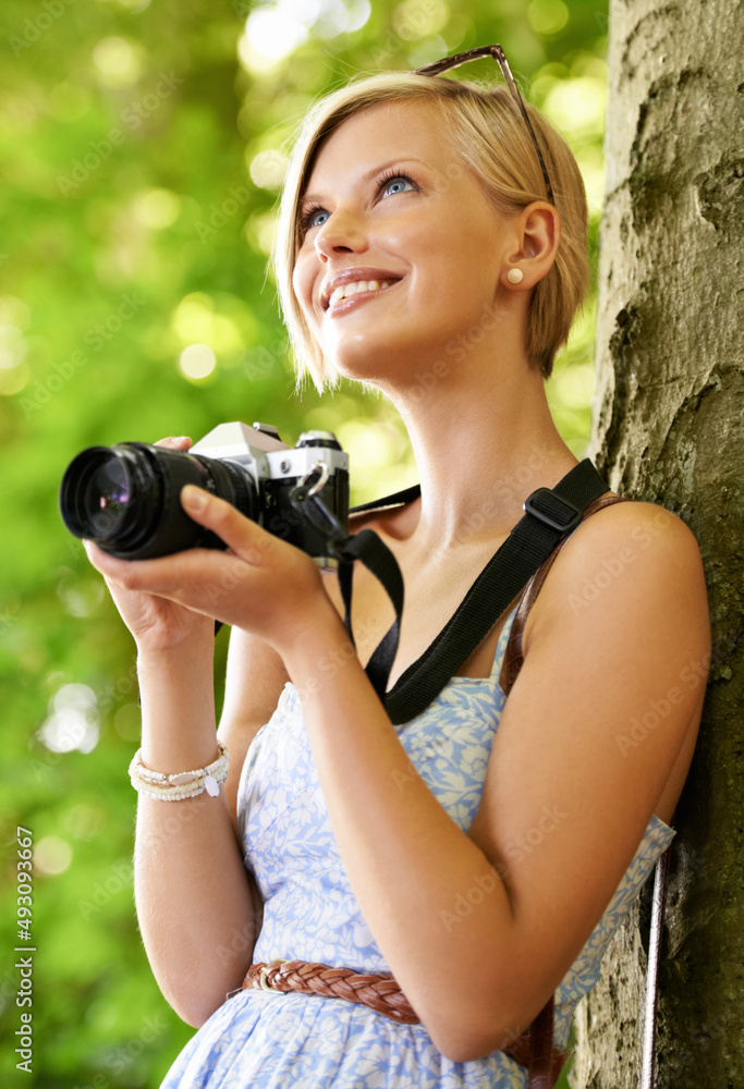 Enjoying taking some photos. Cute young photographer holding her camera and leaning against a tree.