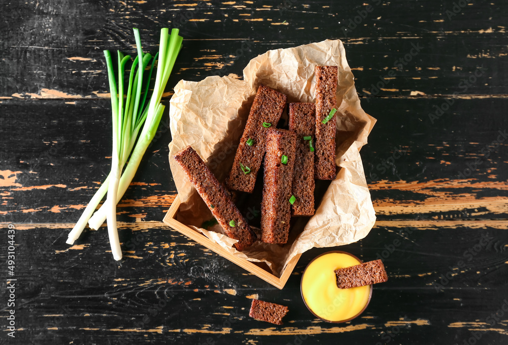 Box with tasty rye croutons and sauce on black wooden background