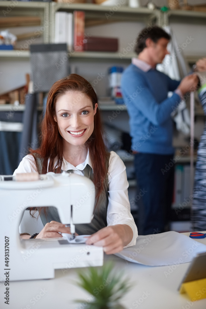 Stitching it all together. Portrait of a young fashion designer sewing while a colleague works in th