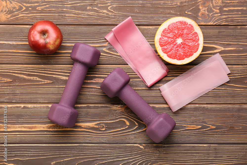 Stylish dumbbells, fruits and elastic bands on wooden background