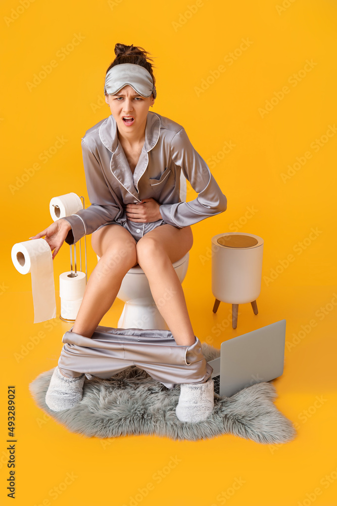 Young woman in pajamas suffering from diarrhea while sitting on toilet bowl against color background