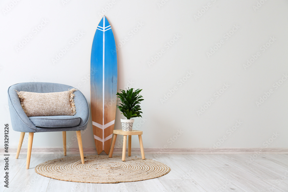 Armchair, stool with houseplant and surfboard near light wall