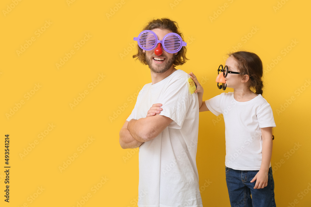 Little girl playing a prank on her father against yellow background. April Fools Day celebration