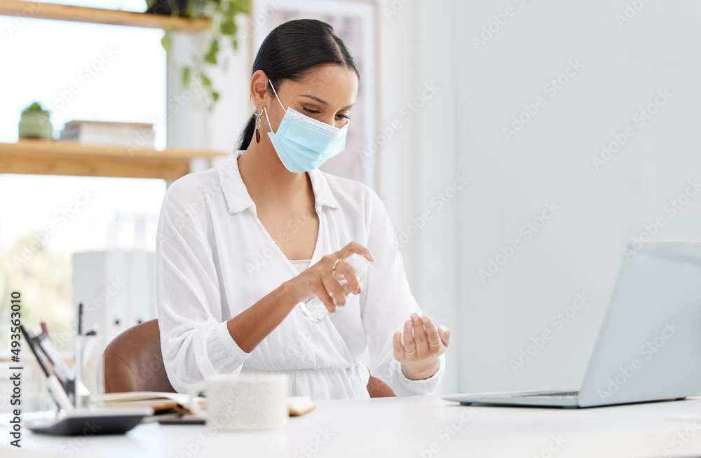 Always staying safe. Shot of a young businesswoman wearing a mask and sitting at a desk sanitising h