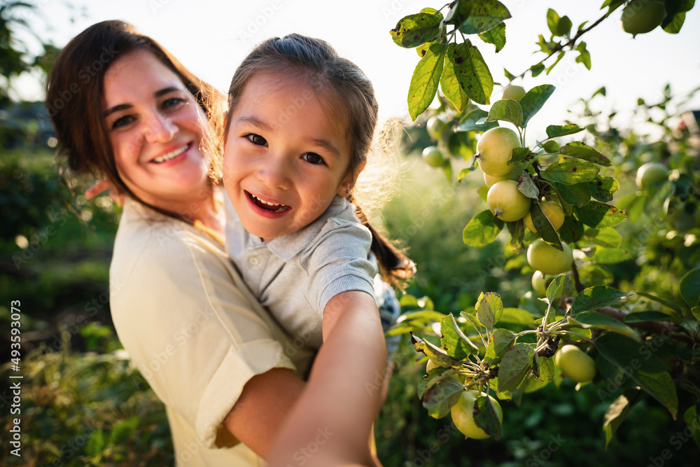 Caucasian mother and her Asian daughter take a selfie in the orchard in summer