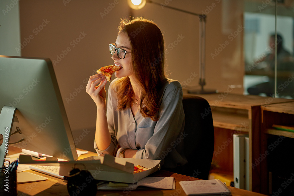 Late night meals in the office. Shot of an attractive young businesswoman eating pizza while working