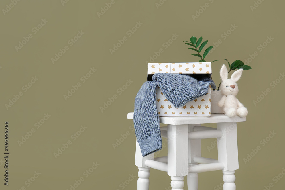 Box with knitted sweater for child and toy on stool against color background