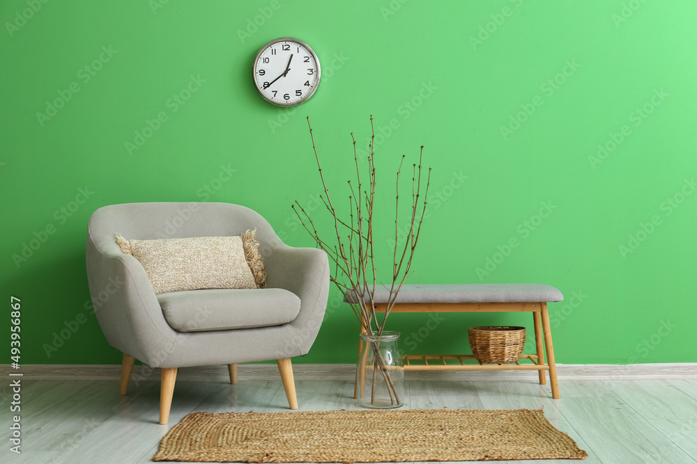 Armchair, vase with tree branches, bench and clock hanging on green wall