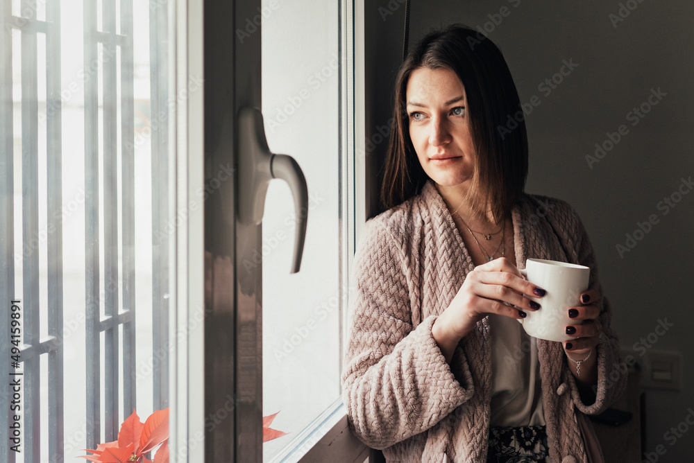 放松的女人一边拿着一杯咖啡，一边看着家里的窗户。