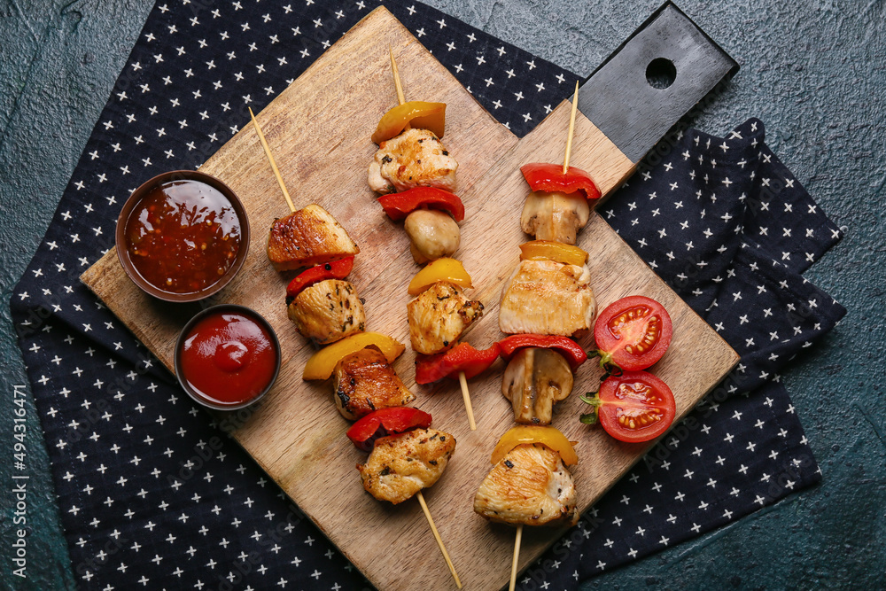 Wooden board of grilled chicken skewers with vegetables and sauces on black background