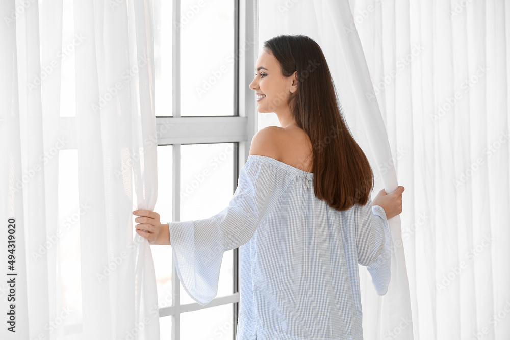 Pretty young woman opening curtains in room