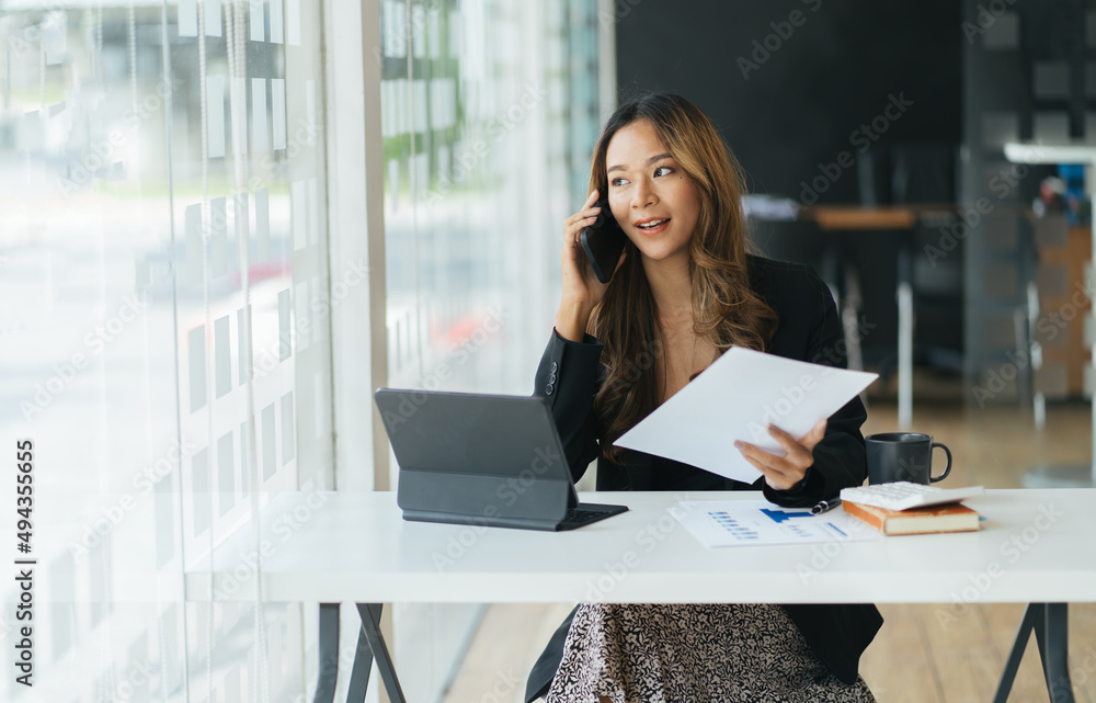 快乐微笑的亚洲女商人在办公室用笔记本电脑工作，使用智能手机。女商人坐着