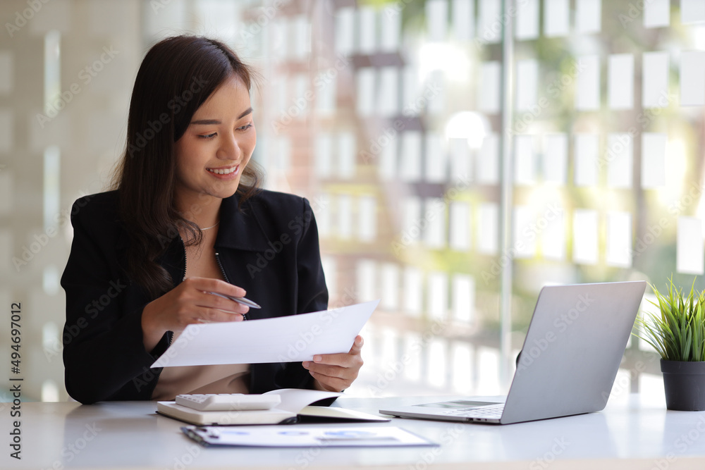 Smiling Asian businesswoman working on laptop and reading documents on desk in office.