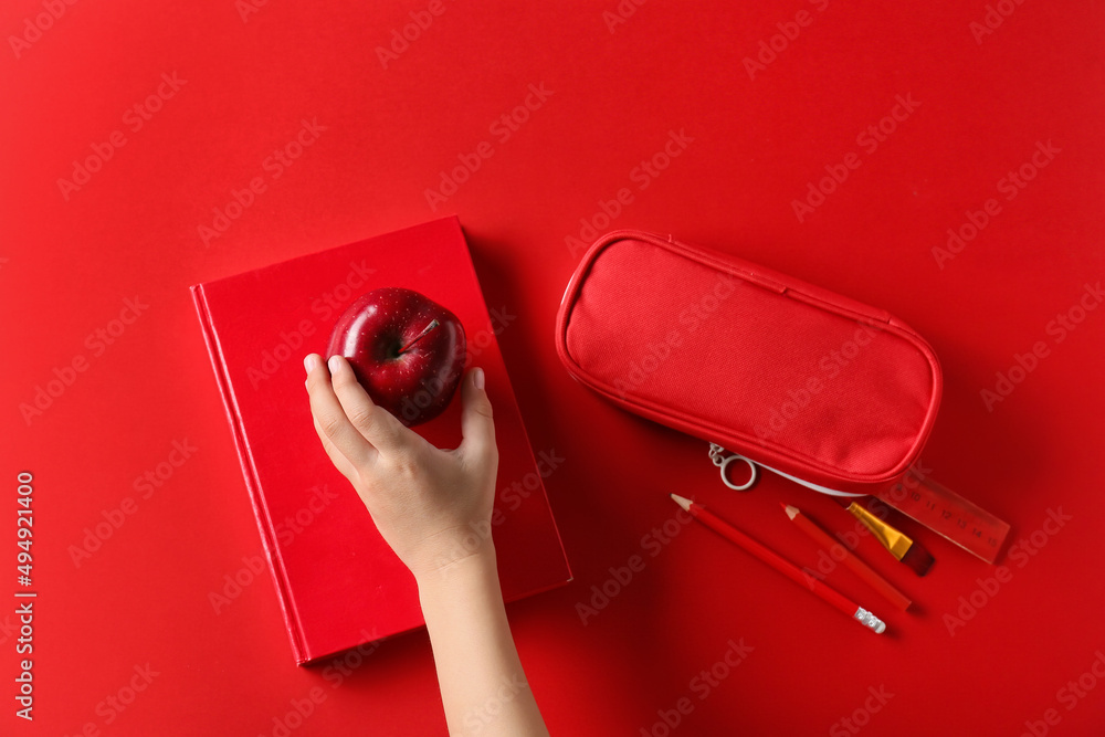 Childs hand with apple, book and pencil case on red background