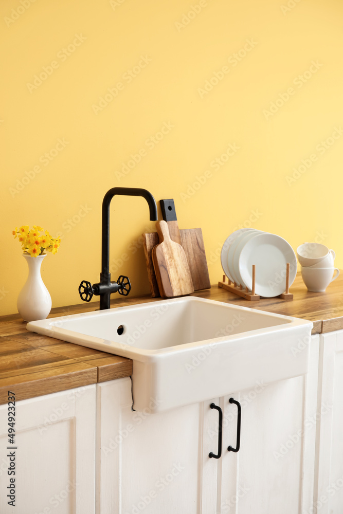 White sink, vase with flowers and kitchen utensils on counters near yellow wall