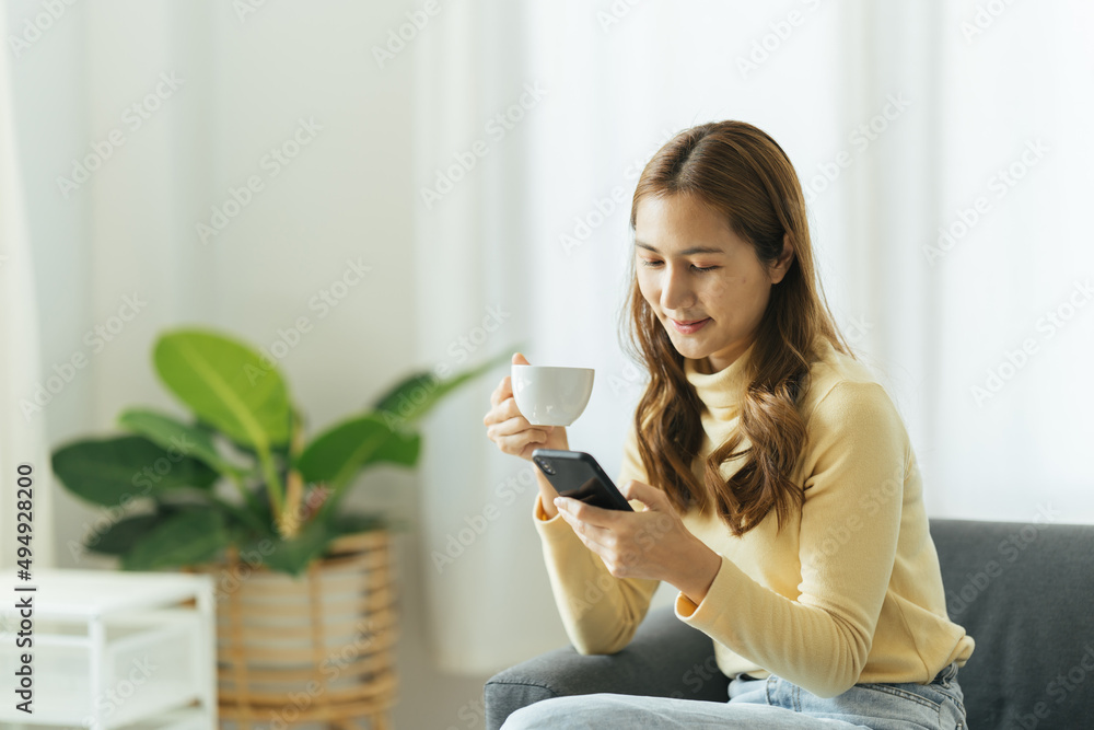 Young beautiful Asian woman relaxing in living room at home, reading books, drinking coffee and usin