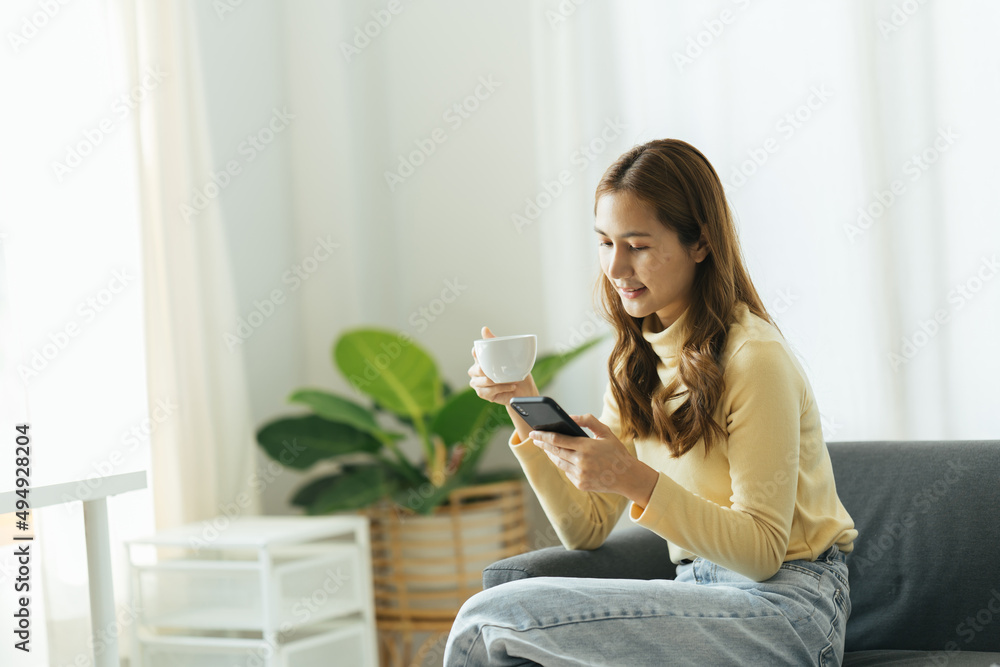 Young beautiful Asian woman relaxing in living room at home, reading books, drinking coffee and usin