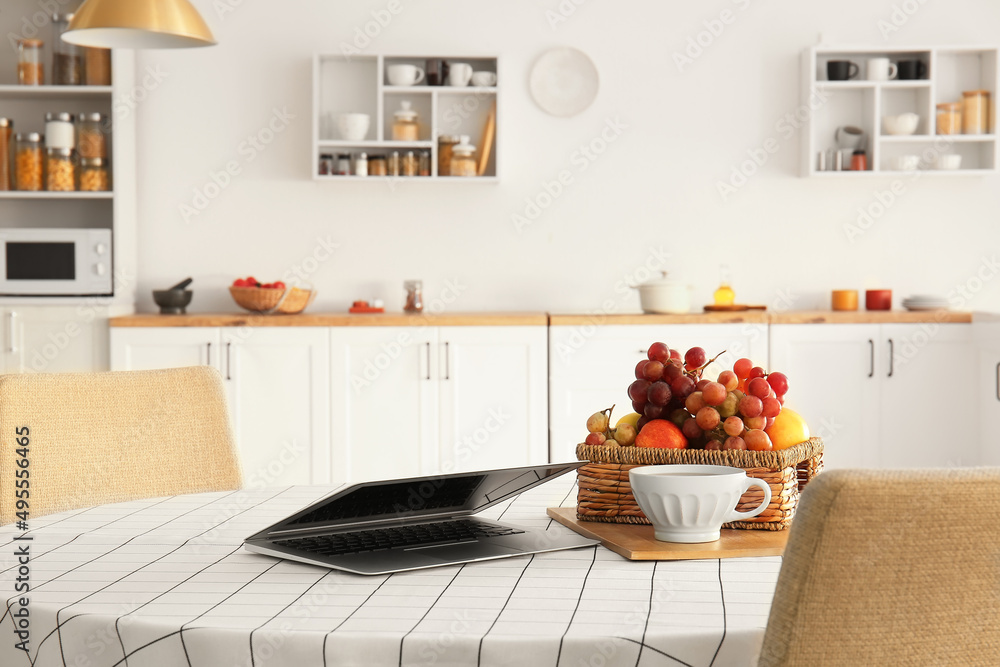 Laptop, cup and fruit basket on dining table in kitchen