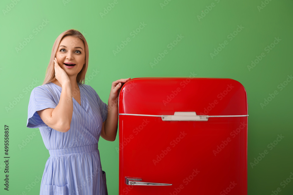 Emotional woman near stylish red fridge on green background