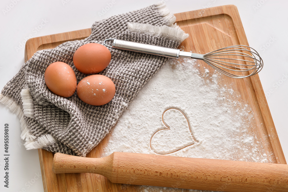 Wooden board with chicken eggs, flour, drawn heart and whisk on light background