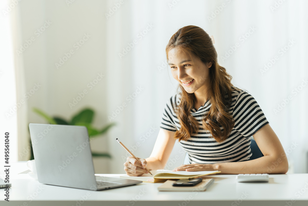 年轻女性在家里的办公室里用笔记本电脑做笔记。