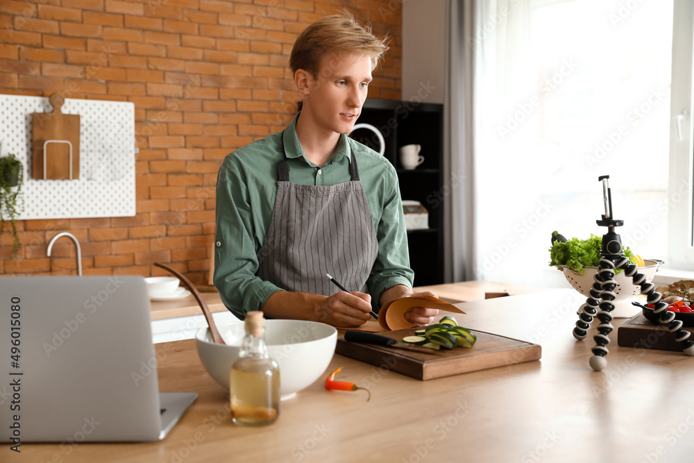 Young man making notes from cooking video tutorial in kitchen