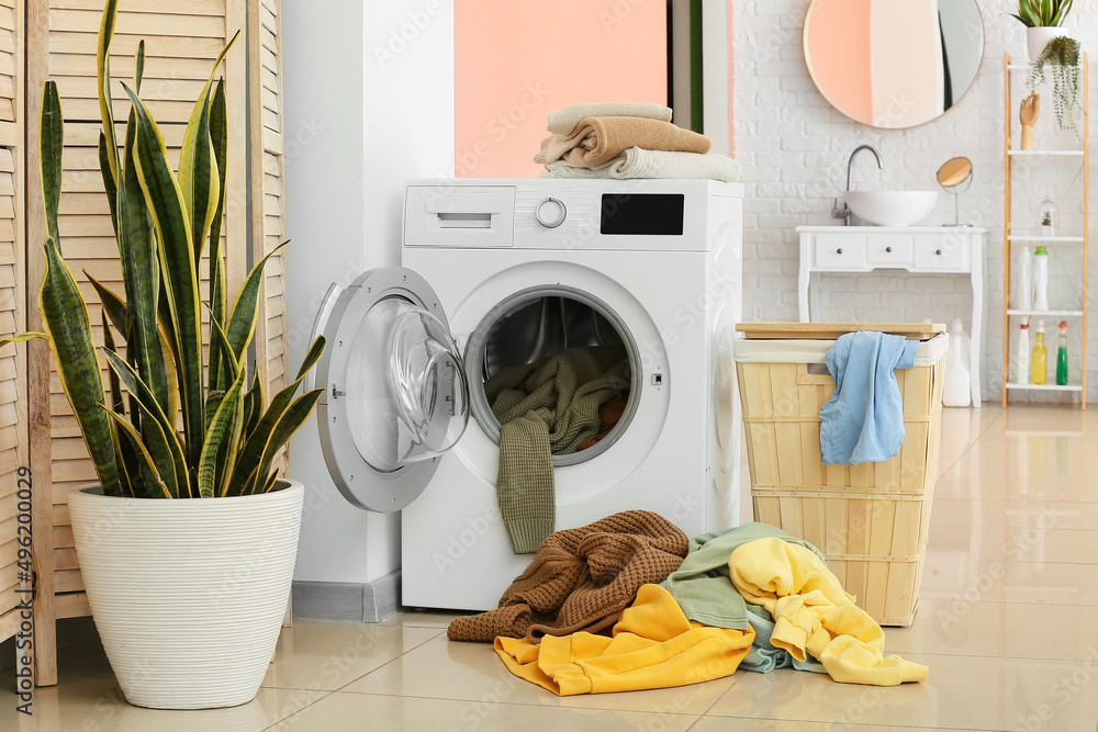 Modern washing machine with laundry, basket and plant in bathroom