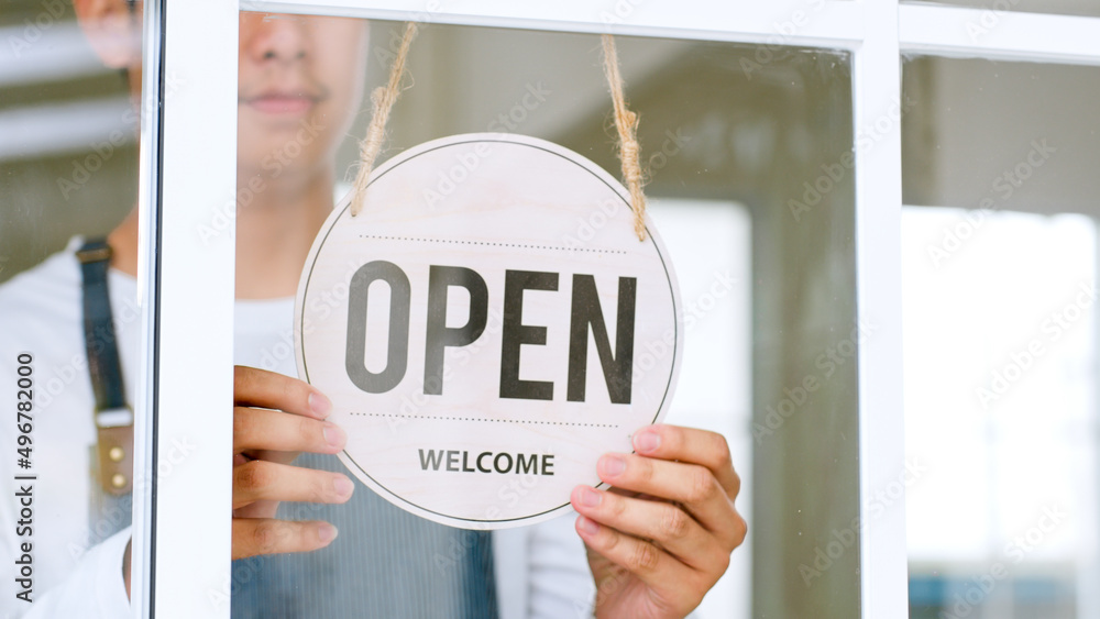 Asian owner in apron turning open sign board on glass door at café, shop, restaurant, small business