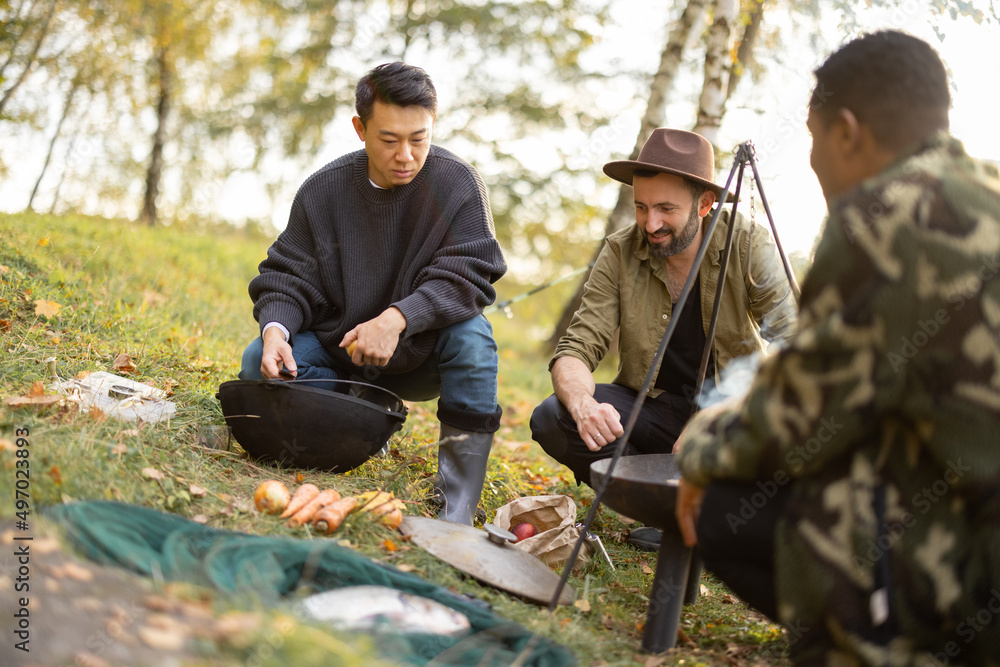 三个多种族的朋友在野餐时用篝火在大锅里煮乌卡汤。男人们在钓鱼和