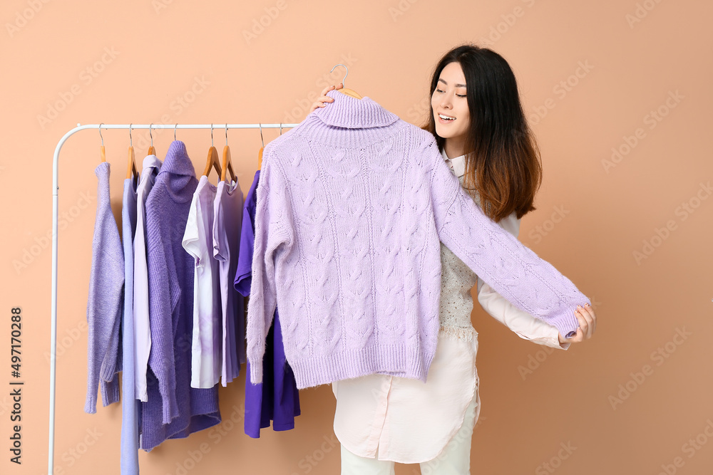 Young Asian woman and rack with clothes in purple shades on beige background