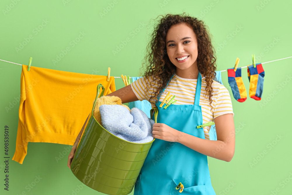 Young African-American woman with laundry basket and clothespins on green background