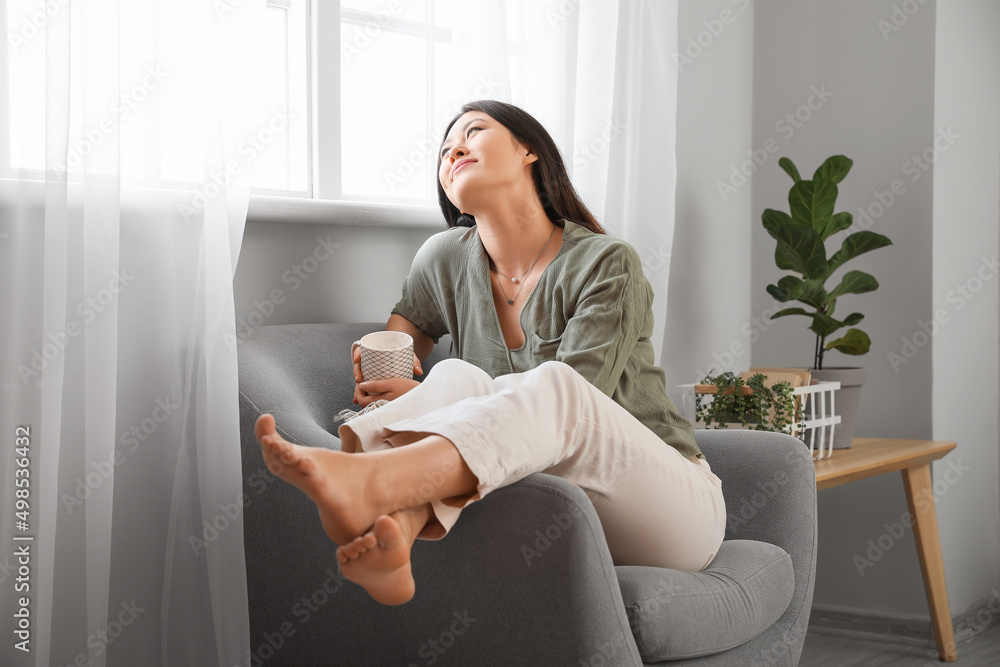 Young Asian barefoot woman with cup of tea sitting in armchair at home