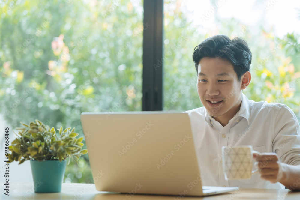 一名身穿白衬衫的亚洲男子拿着咖啡杯，一边用笔记本电脑微笑。