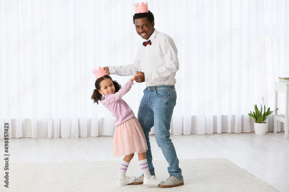 Happy African-American man and his little daughter in paper crowns  dancing at home