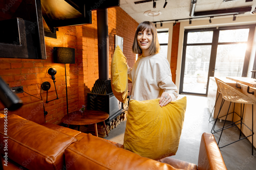 Portrait of a young and pretty woman puts yellow pillows on a couch, while doing some housework at h