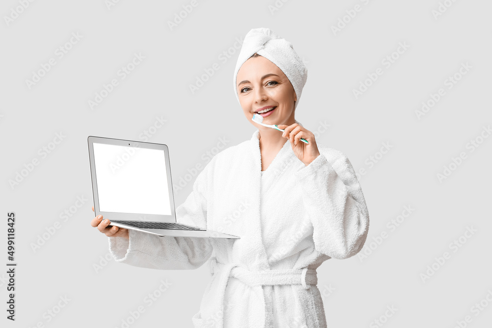 Beautiful mature woman with laptop brushing teeth on light background