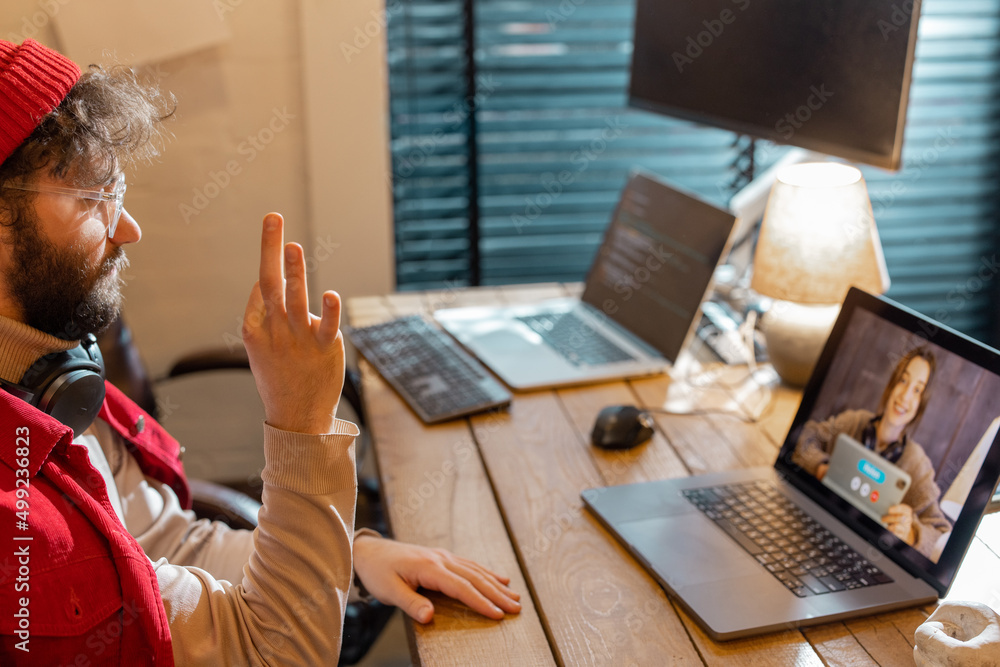 Stylish man in red hat having video call with female colleague while sitting at cozy home office. Co