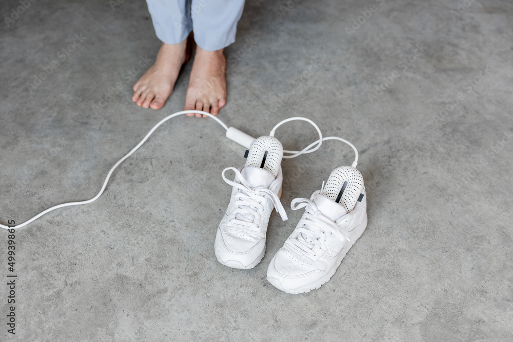 Woman stands barefoot near white sneakers drying with electric shoe dryer on the floor. Concept of m