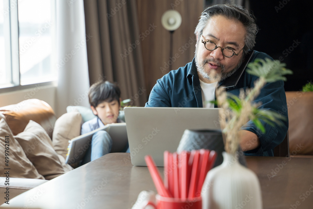 Middle-aged Asian man using laptop work from home with son draws a picture on the back.