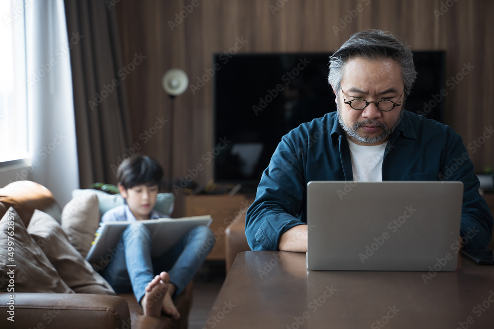 一名亚洲中年男子带着儿子在家用笔记本电脑工作，在背面画了一幅画。