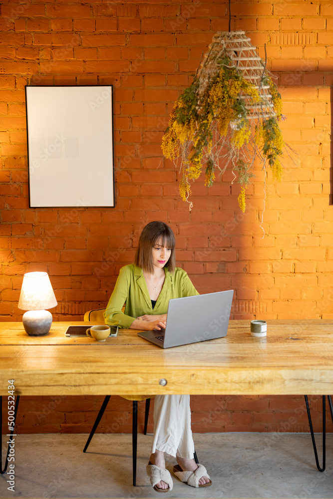 Young elegant woman works on laptop while sitting by the wooden table at cozy home. Remote work from