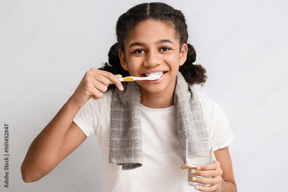 African-American teenage girl with glass of water brushing teeth on light background