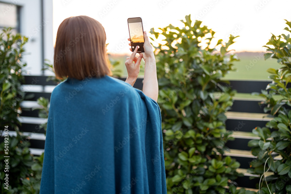 Woman in plaid takes photo on phone, enjoying beautiful sunset at backyard of her country house. Spe