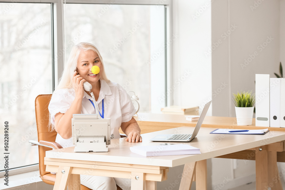 滑稽的成熟女人，带着小丑鼻子在办公室打电话。愚人节庆祝活动