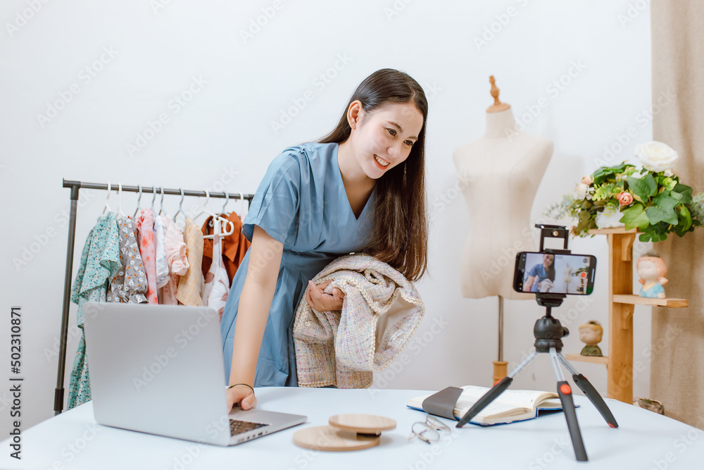 Beautiful Asia woman Live streaming selling clothes online with smartphone at her shop. Small busine