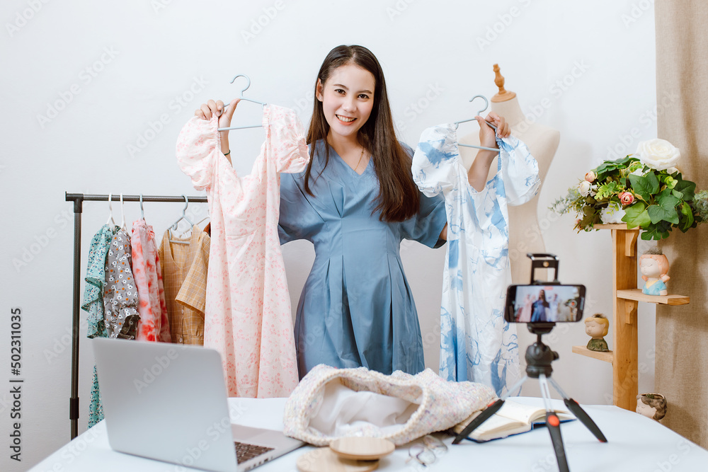 Beautiful Asia woman Live streaming selling clothes online with smartphone at her shop. Small busine