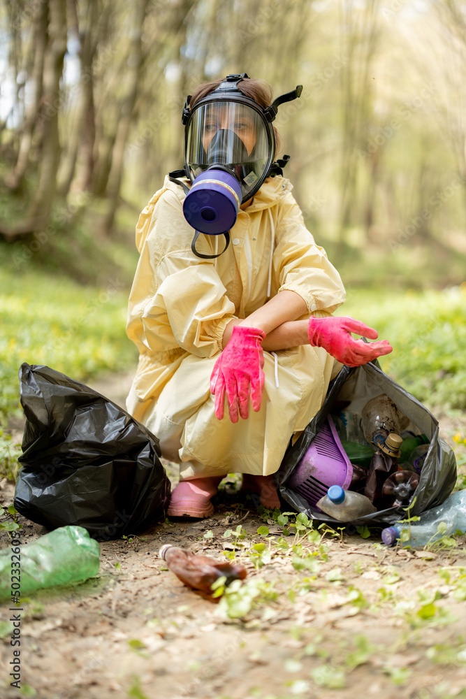 Tired woman in gas mask and protective clothes collecting scattered plastic garbage in the woods. Pr