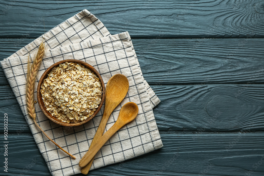 Bowl of raw oatmeal, spoons and spikelets on dark wooden background