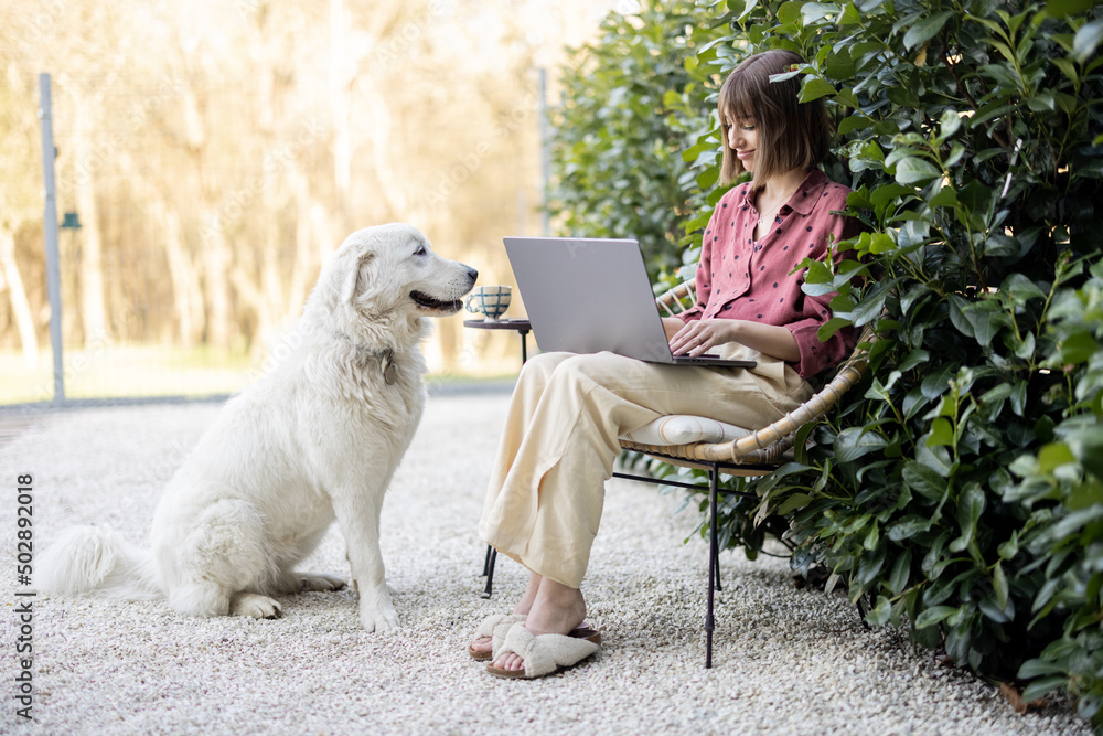 Young woman works on laptop computer while sitting relaxed on chair with her cute adorable dog at ba